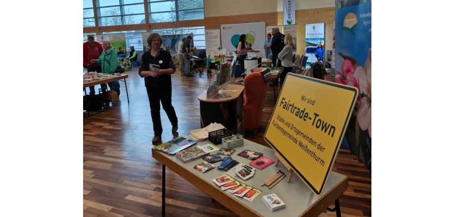 Rechts im Bild der Stand der VG mit einem großen, gelben Schild Fair trade. Im Hintergrund der Blick in die Halle. 