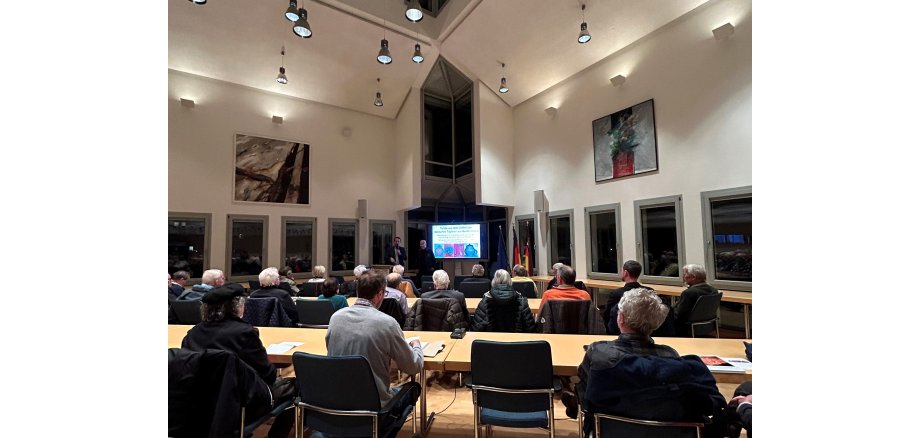 Blick in den Ratssaal mit dem Publikum mit dem Rücken zum Betrachter, ganz vorne der Bildschirm mit der Präsentation sowie dem Referent und dem Bürgermeister.