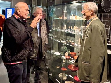 Vor einer Vitrine mit Exponaten steht rechts der Inhaber der örtlichen Tonwerke, auf der anderen Seite der Vitrine Lokalhistoriker Henrichs, dazwischen seitlich vor der Vitrine Referent Urmersbach, der mit der rechten Hand gestikuliert. 