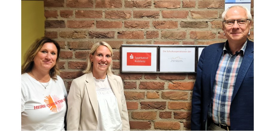 Neben einem Schild mit dem Logo der Sparkasse und der Realschule plus an der Römerville steht rechts Schulleiter Gerhard Müller, links die Ausbildungsleiterin der Sparkasse, Tanja Hommen und ganz links Jobfüxin Katrin Lyding.