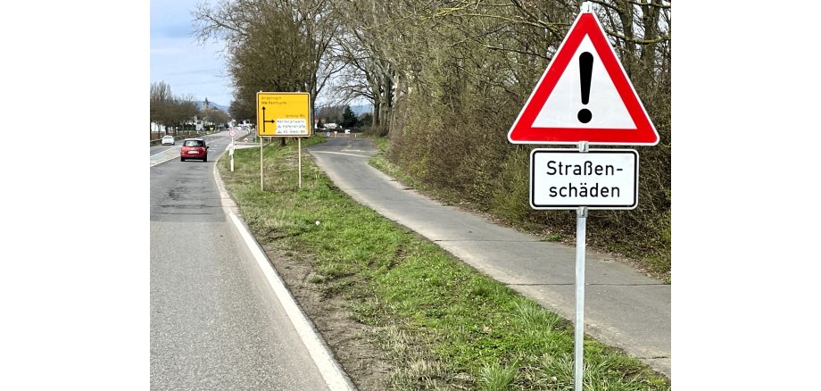 Rechts im Vordergrund ist das dreieckige, rot umrandete Verkehrsschild mit Ausrufezeichen und darunter die Tafel mit der Aufschrift Straßenschäden zu sehen. Im Hintergrund ist die marode Straße.