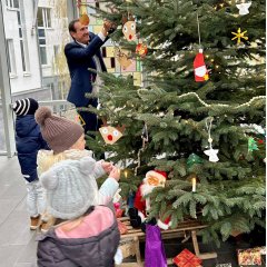 Im Vordergrund stehen drei Kinder in Jacken und mit Mützen am Weihnachtsbaum und schmücken, im Hintergrund Bürgermeister Przybylla, der auch einen Anhänger an den Baum hängt.