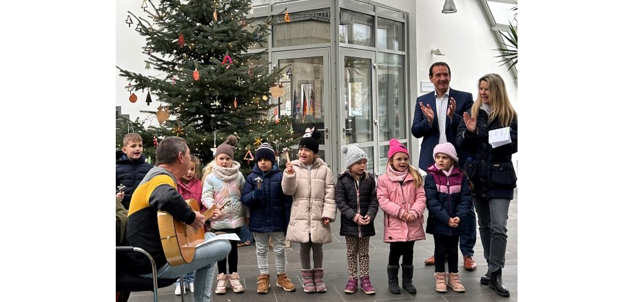 Vor dem Christbaum im Foyer des Rathauses stehen acht Kinder. Links vor ihnen sitzt Erzieher Dirk Bitz auf einem Stuhl und spielt Gitarre. Rechts neben den Kindern steht Erzieherin Ulrike Dinnebier und Bürgermeister Thomas Przybylla.