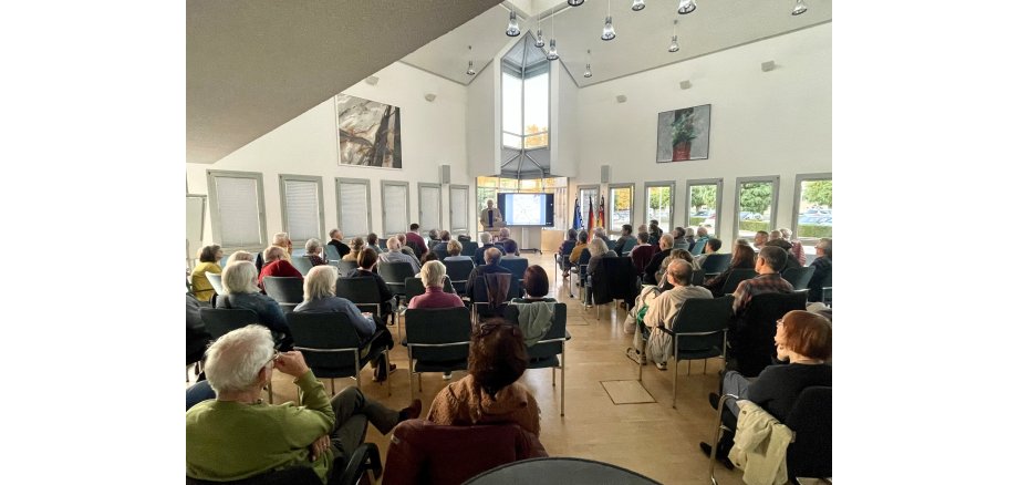 Im Ratssaal der VG sitzt Publikum in zwei Blöcken mit einem Gang in der Mitte. Die Gäste sind von hinten zu sehen. Vorne steht der Referent Dr. Hans-Helmut Wegner neben einer großen digitalen Tafel, auf der eine Präsentation zu sehen ist.