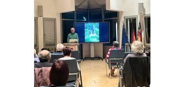 Der Referent Horst Walter steht an einem Rednerpult links neben einer digitalen Tafel, auf der ein Weg und ein Baum zu sehen sind. Im Vordergrund sieht man von hinten Publikum, das in zwei Blöcken links und rechts eines Mittelganges sitzt.