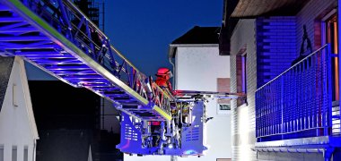 Am linken Bildrand ist eine Drehleiter zu sehen, rechts ist ein Balkon. Die Szene ist in der Dämmerung am frühen Abend. 