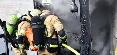 Zwei Feuerwehrmänner sind von hinten zu sehen, wie sie mit einem Schlauch in der Hand einen Brand bekämpfen. Rechts am Bildrand sind verkohlte schwarze Reste und eine unverputzte Wand zu erkennen. 