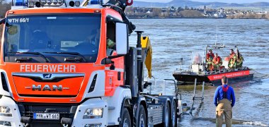 Auf einem Einsatzboot fahren fünf Feuerwehrleute Richtung Rheinufer, wo ein weiterer Feuerwehrmann und ein Feuerwehrfahrzeug warten. Im Hintergrund ist die Silhouette der Stadt Weißenthurm zu erkennen.