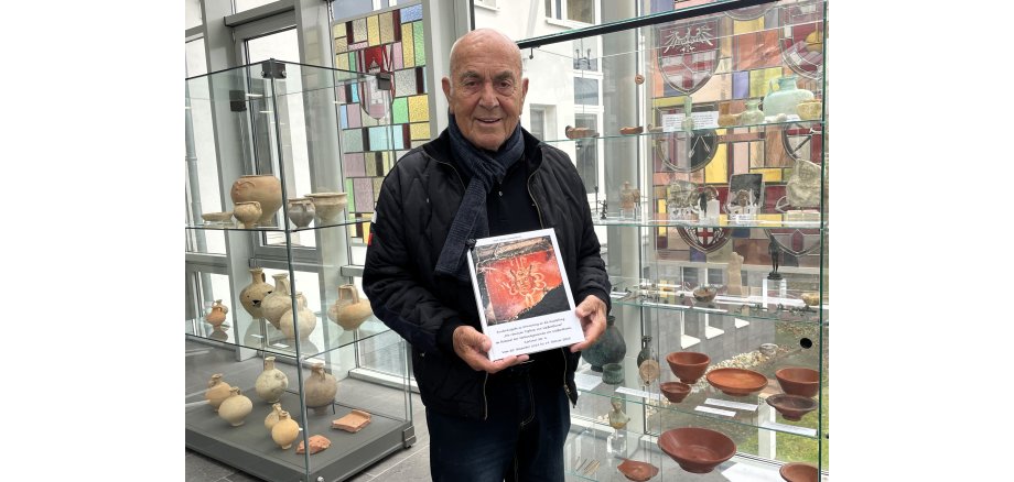 Vor zwei Vitrinen mit Exponaten steht Karl-Heinz Urmersbach und hält sein Buch zu der Ausstellung vor dem Körper.