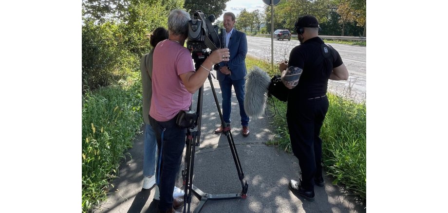 Ein Kameramann des SWR steht in der Mitte, rechts ein Tontechniker, links die Redakteurin, alle mit dem Rücken zum Betrachter. Gegenüber steht Bürgermeister Thomas Przybylla und gestikuliert, im Hintergrund die Straße.