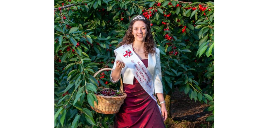die Kirschblütenkönigin Marina I. steht in einem dunkelroten Kleid mit Krone und weißer Schärpe vor einem Kirschbaum, an dem Früchte hängen. An ihrem rechten Arm hat sie einen mit Kirschen gefüllten Korb.