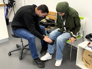 Ein Orthopädietechniker sitzt links auf einem Drehhocker und passt einer Schülerin, die rechts auf einem Stuhl sitzt, eine  Manschette unterhalb des rechten Knies an. 