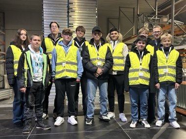 In einer Produktionshalle steht eine Gruppe von 11 Schülerinnen und Schülern, die gelbe Warnwesten und Namensbänder um den Hals tragen, neben Schwerlastregalen.