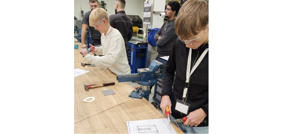 An einer Werkbank stehen stehen zwei Schüler, die Schutzbrillen tragen, und hantieren mit Zangen. Vor ihnen liegt jeweils ein Plan und weiteres Werkzeug, hinter ihnen sind Mitarbeiter des Betriebs und kontrollieren, was die beiden tun.