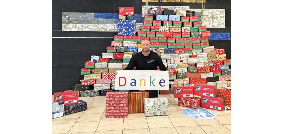 Uwe Fink kniet vor einem großen Stapel Päckchen und hat ein Schild in der Hand, auf dem in bunten Buchstaben "Danke" steht