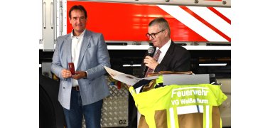 Bürgermeister Thomas Przybylla hält eine Ehrennadel in der Schmuckschachtel in der Hand. Rechts neben ihm steht neben einem Rednerpult Landrat Dr. Alexander Saftig und verliest eine Urkunde. Im Hintergrund ist der Ausschnitt eines Feuerwehrfahrzeugs zu erkennen. 