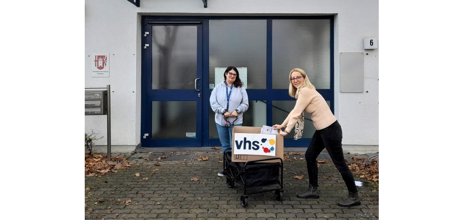 Vor der blauen Eingangstüre in ein Gebäude stehen links und rechts eines Bollerwagens zwei Mitarbeiterinnen der Volkshochschule. Die FRau rechts hält ein Schild mit dem VHS-Logo, das auf dem Bollerwagen liegt. 