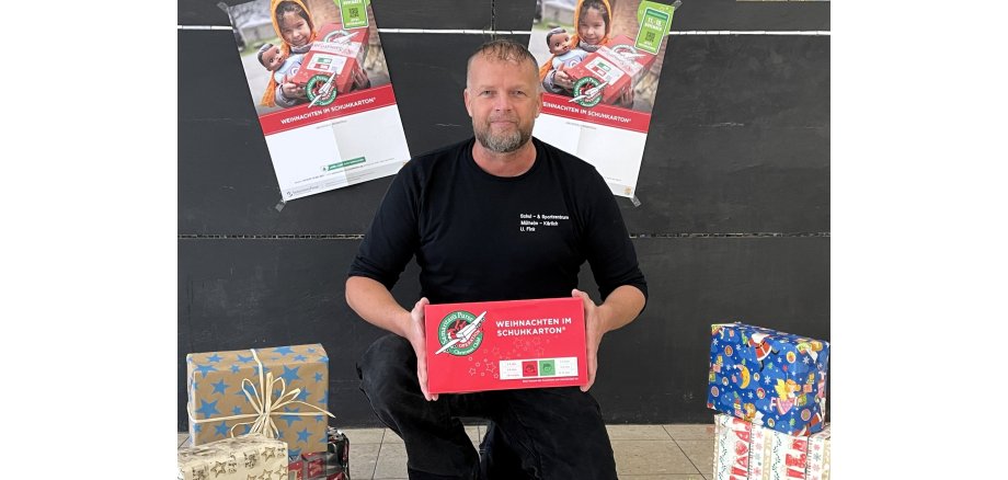 Hausmeister Uwe Fink kniet am Boden neben w Päckchen in Weihnachtspapier und hält ein Paket der Aktion in der Hand. Hinter ihm hängen zwei Plakate von Weihnachten im Schuhkarton an der Wand. 