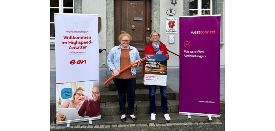 Die Bassenheimer Bürgermeisterin Natalja Kronenberg steht mit einer Vertreterin der Firma Westconnect vor dem Bassenheimer Rathaus. Links und rechts von den beiden stehen Banner der Firmen Westconnect und eon. Frau Kronenberg hat ein Stück Glasfaserkabel in der Hand, die Westconnect-Mitarbeiterin hat ein Werbeplakat für schnelles Internet in der Hand.