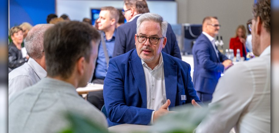 Dozent Jörg Eisbach sitzt mit  weißem Hemd und blauem Jacket zwischen Businessleuten und erklärt etwas. Im Vordergrund sind zwei Männer in weißen Hemden von hinten zu sehen, im Hintergrund stehen Grüppchen zusammen und reden miteinander. 