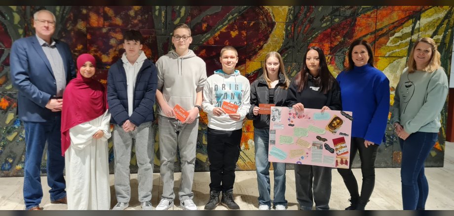 Die Preisträgerinnen  und -träger stehen mit Schulleiter Müller, Jobfüxin Katrin Lüding und Wirtschaftsförderin Kristina Uerz in der Aula der Schule und zeigen ein Plakat.