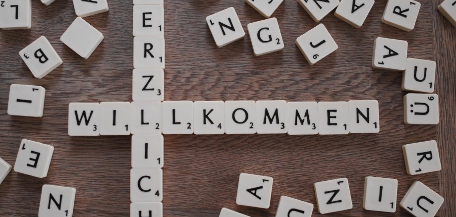 Mit den quadratischen Buchstabenplättchen des Spiels Scrabble ist von oben nach unten das Wort Herzlich und von links nach rechts mit einer Begegnung bei dem L das Wort Willkommen gelegt. Darum herum liegen weitere Plättchen mit anderen Buchstaben.