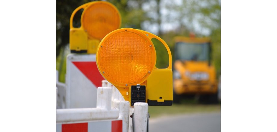 In Nahaufnahme eine Straßenabsperrung mit zwei orangefarbenen Leuchten zu sehen, im Hintergrund Bagger. 